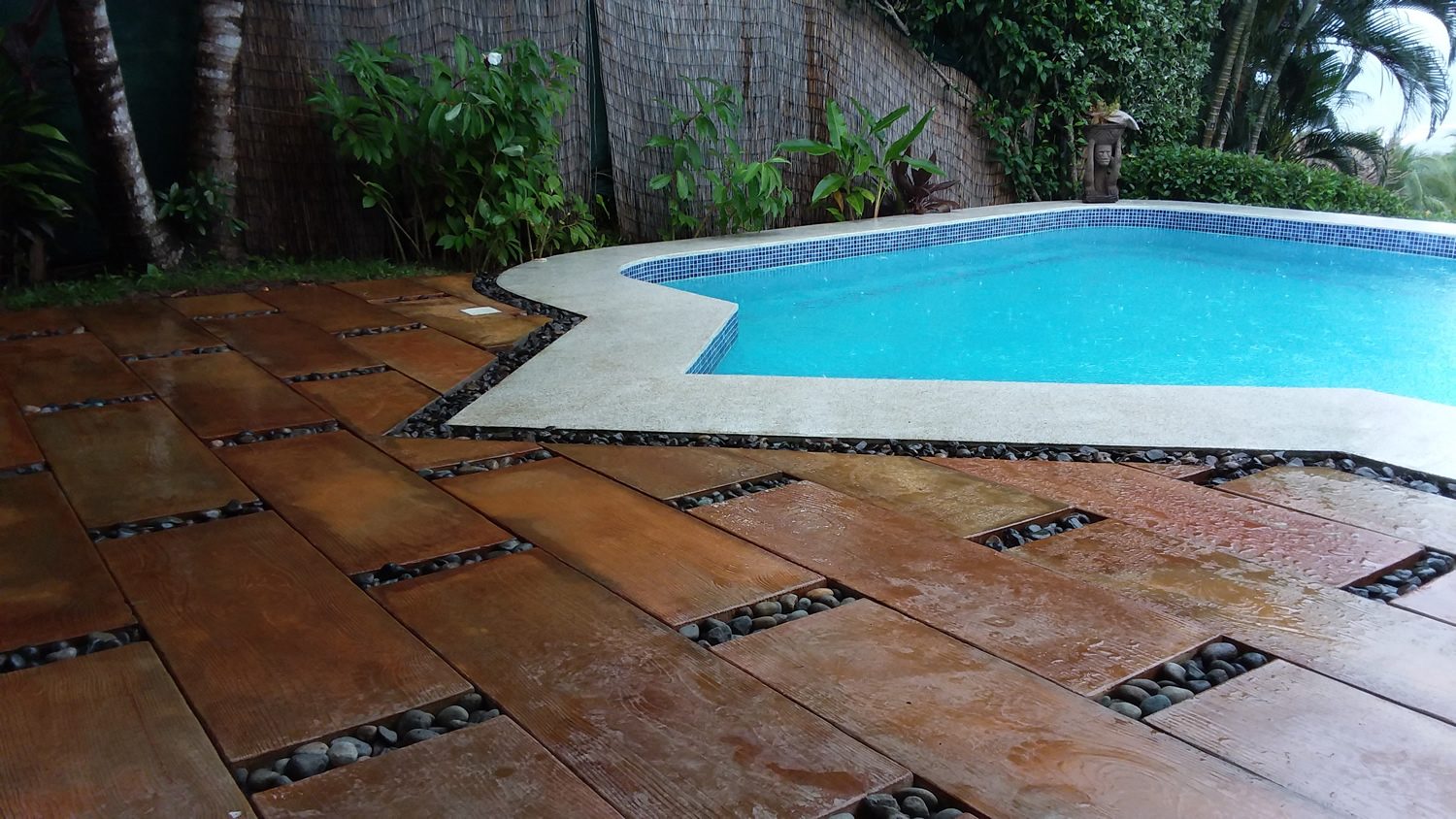 Terraza de piscina en baldosa Madera Grande rojiza, con piedra de río negra en las juntas